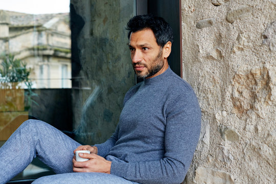 Junger Mann sitzt auf Terrasse vor Fenster mit Spiegelung von Altstadt und Steinwand trinkt Kaffee