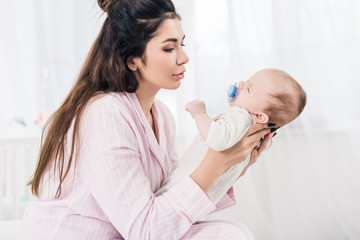 side view of young mother looking at little baby with pacifier at home