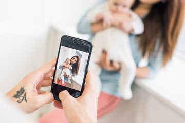 cropped shot of man taking picture of wife with baby in hands on smartphone