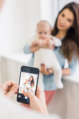 cropped shot of man taking picture of wife with baby in hands on smartphone
