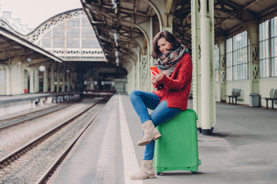 Pleased young woman has positive look, sits crossed legs at bag, holds cell phone, searches place on online map, makes video call, surfes internet website, waits for train at railway station