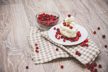 On a light tabletop on a linen napkin in a saucer cake with whipped cream with fresh berries, a bowl with red berries.