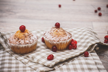 Two cupcakes, sprinkled with powdered sugar and decorated with red berries on a linen checkered napkin on a light wooden table top.