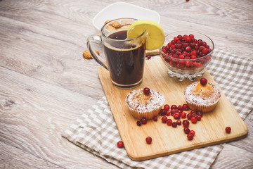 On a light wooden tabletop on a linen napkin napkin, there is a cutting board with two muffins, a broken chocolate bar and bright red berries in a small tree, next to a bowl with cookies.