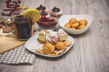 Lemon tea, sesame biscuits in a white bowl, muffins decorated with chocolate, a bowl of red berries, and a plate of sweets are served on a light wooden tabletop.
