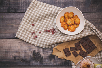 Sesame biscuits in white bowl, dark chocolate broken on craft paper, linen napkin with cranberries on a dark black background of a wooden tabletop