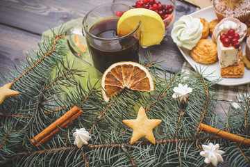 Obraz na płótnie Canvas Close-up of blue spruce twigs, gingerbread cookies, cinnamon sticks, slices of dried orange and white meringues, a glass of tea with lemon and sweets on a white plate — all on a gray wooden table top.