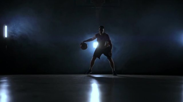 Skill dribbling basketball player in the dark on the basketball court with backlit back in the smoke. Slow motion streetball