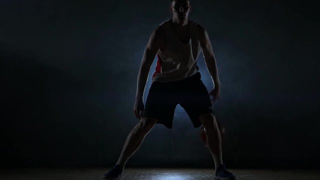 Dribbling basketball player on the court with the ball in a dark room with a backlight in slow motion in the smoke