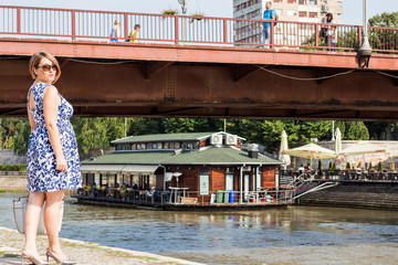 Beautiful Brunette Girl in a Blue Dress Posing Next to the River With River Cafe Boat Behind Her