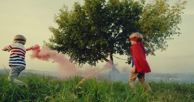 Cute children in the costumes of superheroes running with a wooden model of a plane and playing together in the colorful smoke outdoor at the big tree.