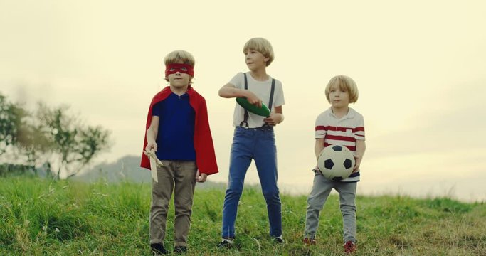 Three cute small teenage boys - one with a football ball, one in the costume of superhero with a sword and one with a frisbee plate playing in front of the camera in the nature.