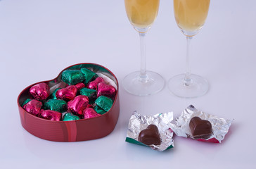 box with chocolates in the shape of a heart, wrapped with fuchsia and green colors and two glasses with champagne, on a white background