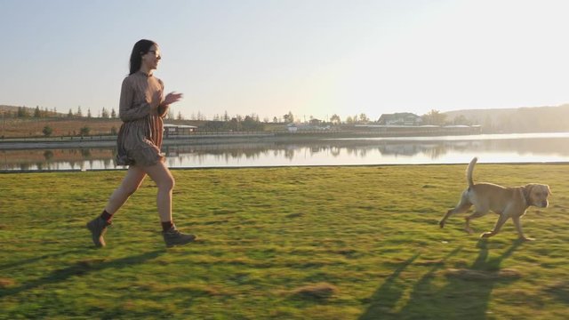 Adorable young woman playing running with her cute dog on nature near the lake.