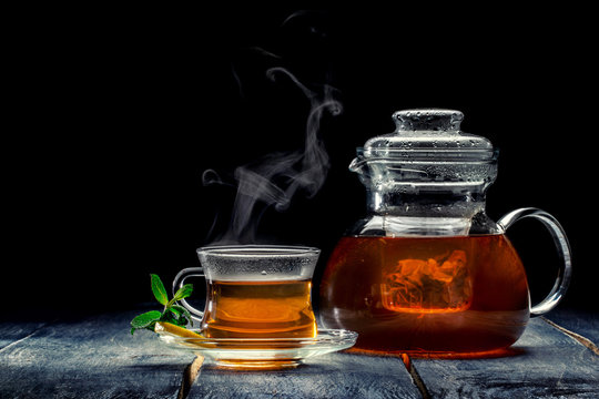 Freshly brewed black tea with mint and lemon in glass cup and glass teapot on a wooden table, on a dark background
