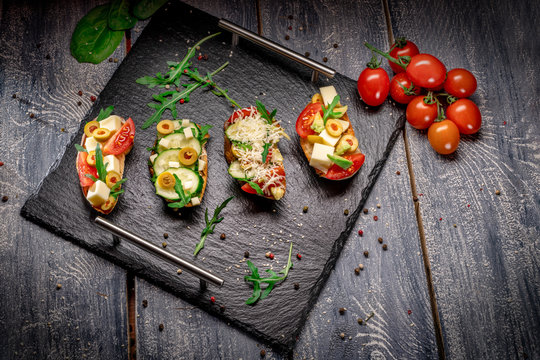 Fresh delicious bruschetti. Variety of small sandwiches with parmesan cheese, cherry tomato, egg, cucumber, avocado on rustic wooden board over dark background