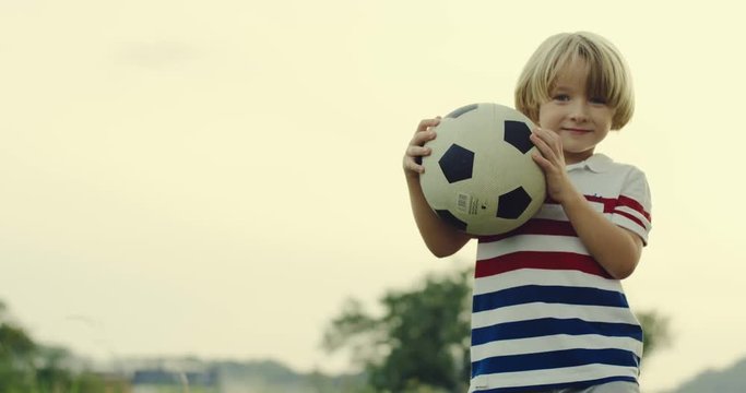 Portrait of the cute small Caucasian boy smiling to the camera with a football ball in hands. Outside.