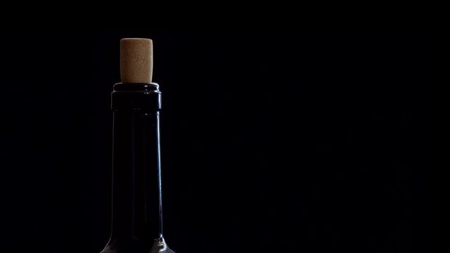 The neck of a wine bottle, with a cork sticking out of the top, is shown, slowly rotating in place, set against a solid black background.