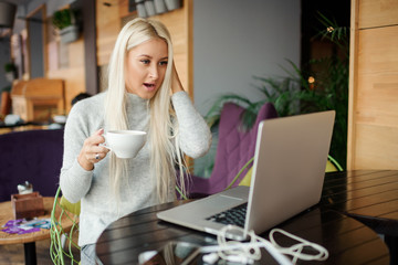 Shocked young woman in casual wear working on laptop while sitting in creative office or cafe and woking. Holding a cup of coffee and smiling 