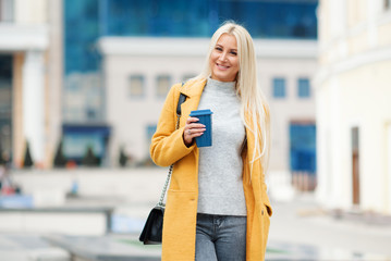 Coffee on the go. Beautiful young blond woman in bright yellow coat holding coffee cup and smiling while walking along the street 