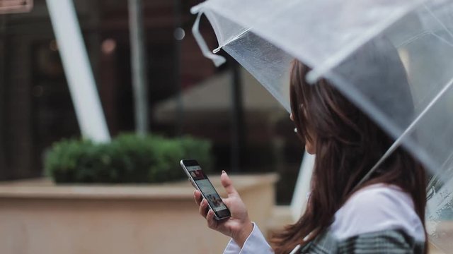 Beautiful young business woman using smartphone walking on the street in rainy weather, smiling, holding umbrella, communication concept