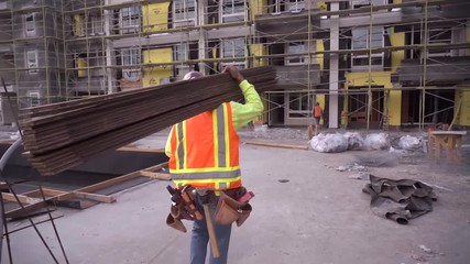 A construction worker walking and working on a construction site wearing a tool belt and high visibility clothing carrying lumber. 