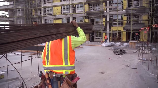 Construction worker walking in slow motion and working on a construction site wearing a tool belt and high visibility clothing carrying lumber. 