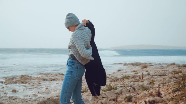 FUNNY: A young woman is trying really hard to put on a jacket in a strong wind. She is on a shore with an ocean in the background