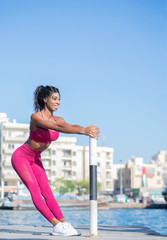 Beautiful tall athletic African American Woman does a stretch pose wearing a bright pink workout outfit with a river and old boats and cityscape in the background on a bright sunny day 