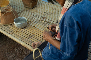 Selective focus on hands of old man makes traditional basket by weaving the natural material.