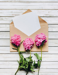 Three faded purple peonies and an open brown kraft envelope containing a white sheet of paper. Valentine's Day concept. Copy space, top view.