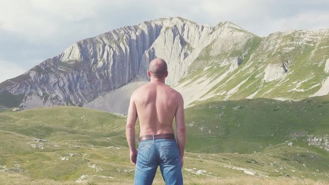 View from back of man looking at mountains. Stock. Man in jeans and shirtless enchanted looks at rock. Courageous and heroic image of man on background of powerful landscape of mountains