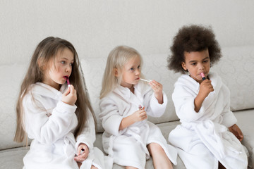 Girlfriends of girls paint lips with lipstick. Three little girls in white coats after a shower playing with makeup paint their lips with red lipstick.