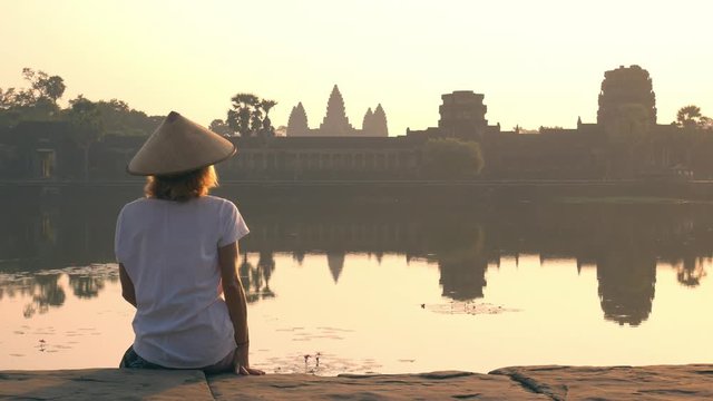 Woman visiting Angkor Wat ruins at sunrise, famous travel destination Cambodia. Reflection on water pond and sunburst, soft light, toned.