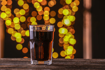 Glass of tasty cola drink on wooden table against blurred lights