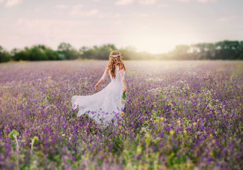 young lady dressed in an elegant long white dress with transparent sleeves and , with a neat hairstyle of blond hair decorated with a white wreath, walks across the field of purple flowers. no face