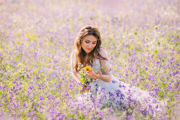 lovely sweet woman with closed eyes dressed in an elegant white dress with transparent sleeves sits in purple flowers with a natural field bouquet in her hands. atmospheric photo, gentle warm colors