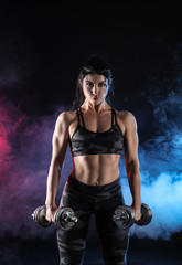 Fototapeta na wymiar Sporty muscular woman with dumbbells in smoke on dark background