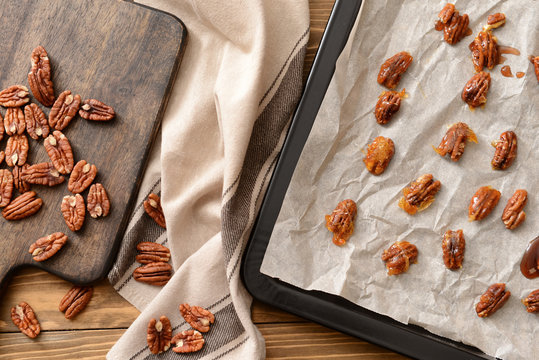 Board and baking sheet with raw and candied pecan nuts on wooden table