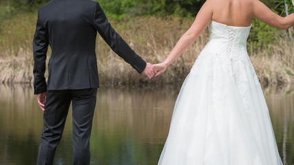 Fototapeta na wymiar Hochzeitspaar in schönen Kleidern steht vor einem Teich und hält sich an den Händen