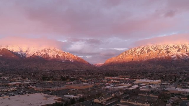 Aerial view flying backwards from snow capped mountains at sunset over city center in Utah Valley.