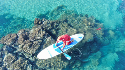 Aerial photo of fit man dressed as Santa Claus practising SUP or Stand Up Paddle in exotic mediterranean beach with turquoise sea