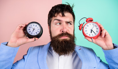 Man bearded hipster hold two different clocks. Guy unshaven puzzled face having problems with...