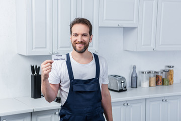 smiling male handyman in working overall showing card with lettering home inspection