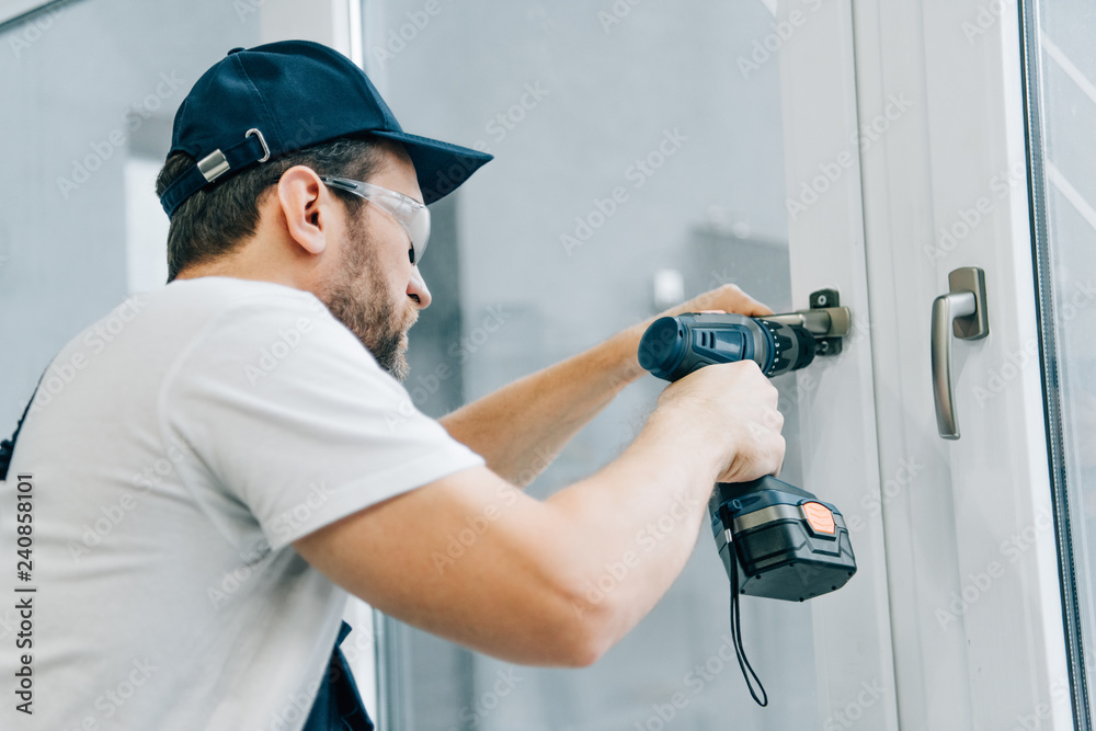 Wall mural handyman in goggles fixing window handle by electric drill