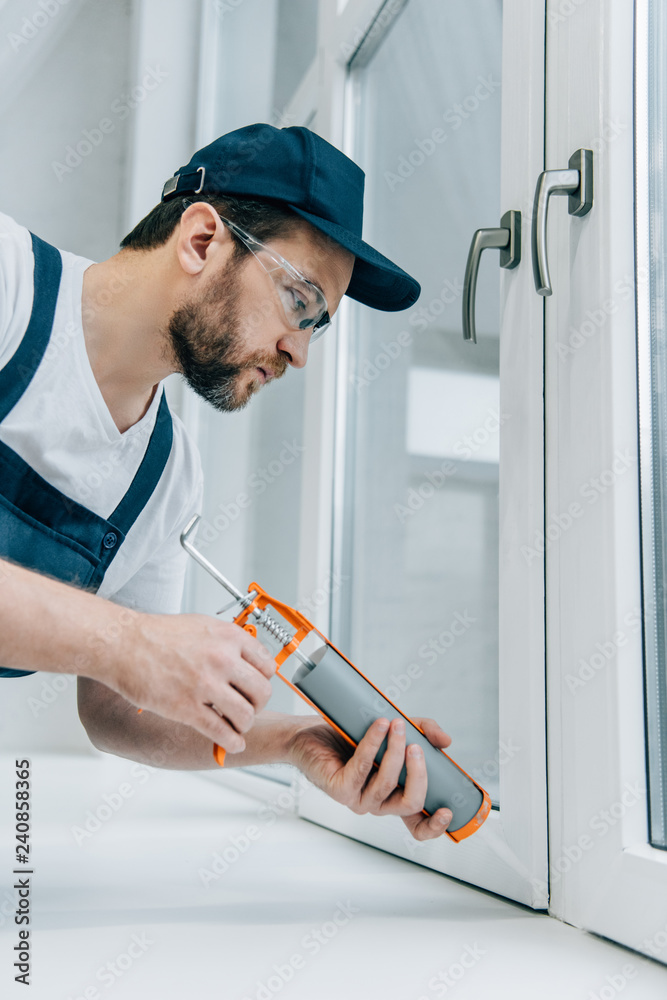 Wall mural side view of adult repairman in goggles fixing window with sealant gun