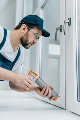 side view of adult repairman in goggles fixing window with sealant gun