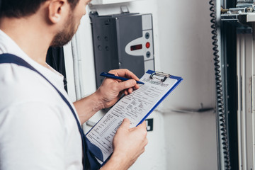 partial view of male electrician writing in clipboard and checking electrical box