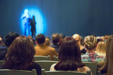 Man appears on stage in theater with many people