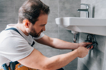 adult male plumber in working overall fixing sink in bathroom